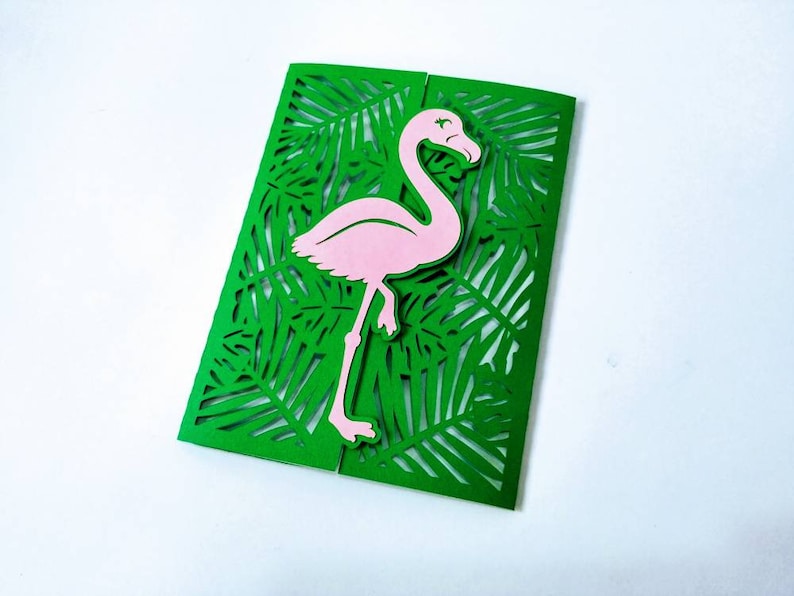 May include: A green card with a pink flamingo cutout. The card is decorated with a laser cut design of tropical leaves.