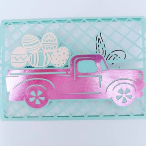 May include: A pink pickup truck with a white bed carrying pastel Easter eggs. The truck is cut out of paper and is sitting on a light blue background.