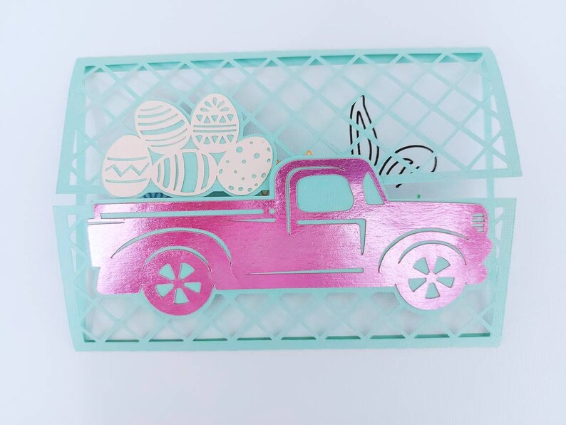 May include: A pink pickup truck with a white bed carrying pastel Easter eggs. The truck is cut out of paper and is sitting on a light blue background.