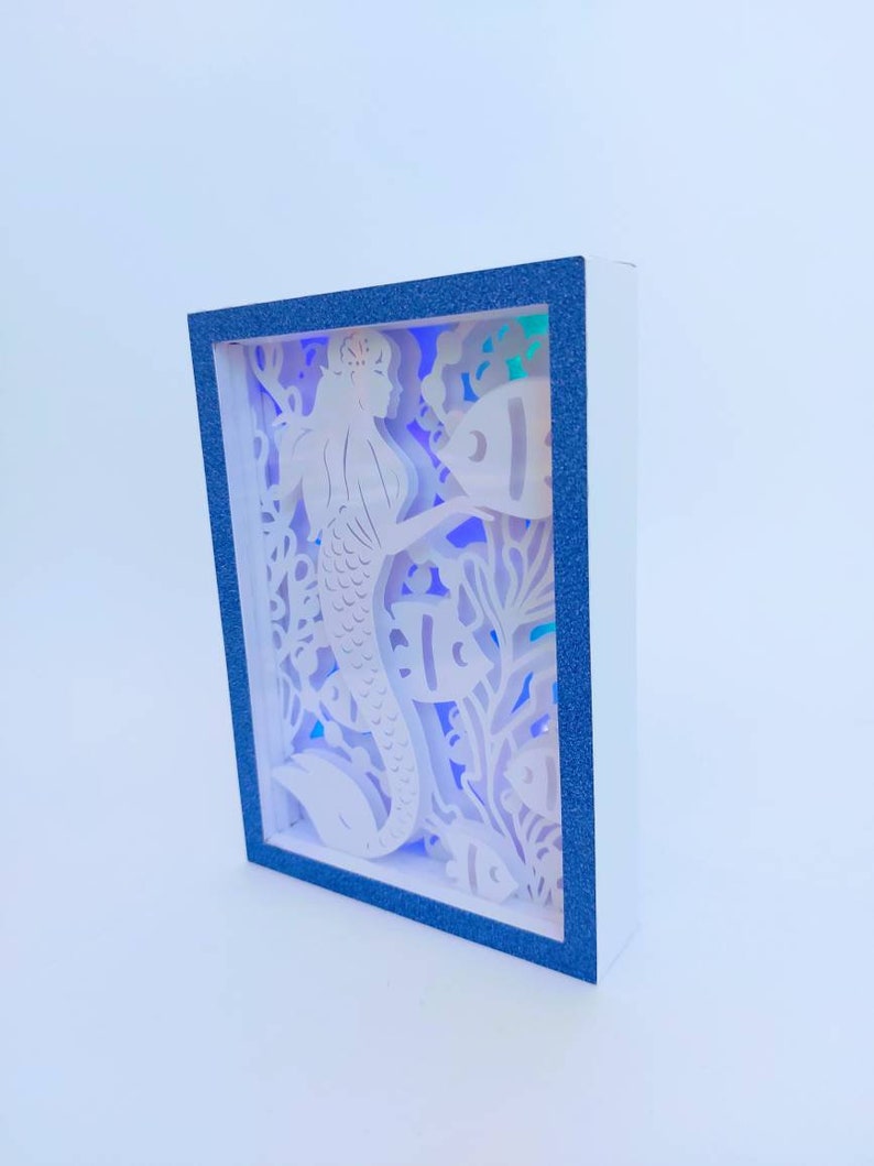 May include: A white papercraft mermaid with a crown and tail, surrounded by fish and coral, is illuminated by a blue and purple light. The papercraft is framed in a blue frame.