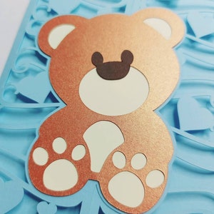 May include: A light blue card with a brown teddy bear cutout. The teddy bear has white paws and a white face with brown eyes and a brown nose. The card is decorated with a blue and white pattern.