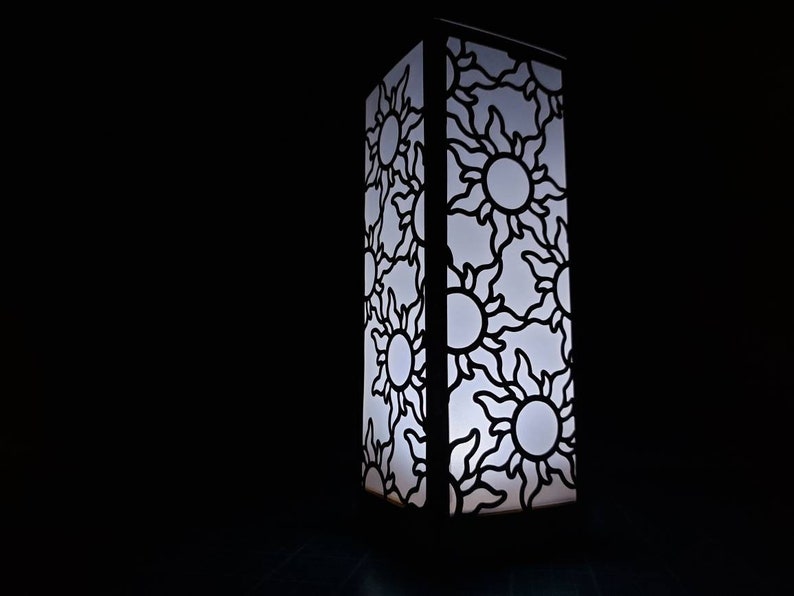 May include: A white lamp with a black cutout design of suns. The lamp is lit and casting a warm glow.