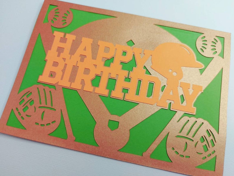 May include: A green and orange paper card with a baseball theme. The card has a baseball diamond, bats, gloves, and baseballs cut out. The words "Happy Birthday" are in orange letters.