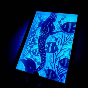 Peut inclure: Une lumière bleue éclaire une découpe en papier blanc d'une sirène avec une queue de poisson et de longs cheveux, entourée de poissons et d'algues. La découpe est encadrée dans un cadre en bois.