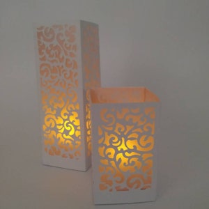 May include: Two white candle holders with intricate cut-out designs. The holders are illuminated from within, creating a warm glow.