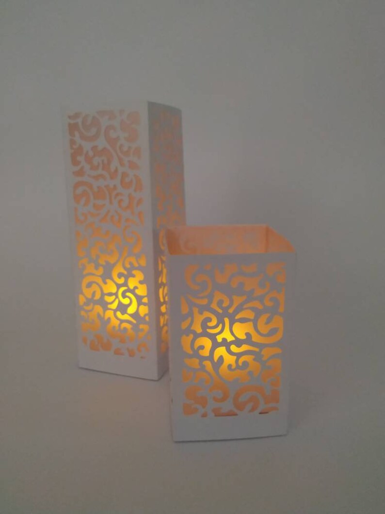 May include: Two white candle holders with intricate cut-out designs. The holders are illuminated from within, creating a warm glow.