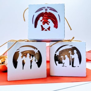 Könnte beinhalten: Weiße, würfelförmige Weihnachtsschmuckstücke mit ausgeschnittenen Krippenszenen. Jedes Ornament zeigt eine andere Szene, darunter die Heilige Familie und die Heiligen Drei Könige. Goldene Schnurschlaufen sind angebracht. Rote und goldene Weihnachtskugeln sind im Hintergrund.