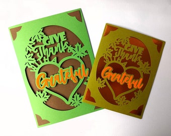 Pode incluir: Dois cartões de felicitações feitos à mão com fundo verde e dourado. Os cartões apresentam um coração castanho com o texto "Give Thanks with a Grateful Heart" em letras laranja. O coração é rodeado por folhas recortadas.