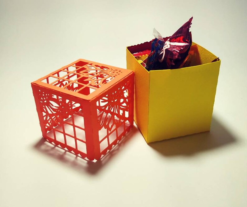 Puede incluir: Dos cajas peque&ntilde;as cuadradas. Una caja es naranja con un dise&ntilde;o recortado y la otra es amarilla. La caja amarilla est&aacute; llena de caramelos rojos y blancos.