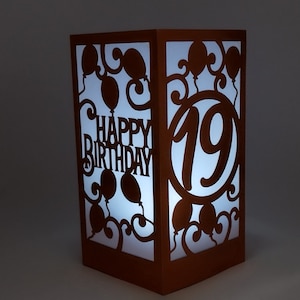 May include: A brown paper lantern with a white light shining through it. The lantern has a cut-out design of balloons and the words "Happy Birthday" and the number "19".