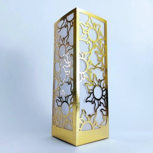 May include: A gold-colored decorative box with a cutout design of stylized suns. The box is made of cardboard and has a white interior.