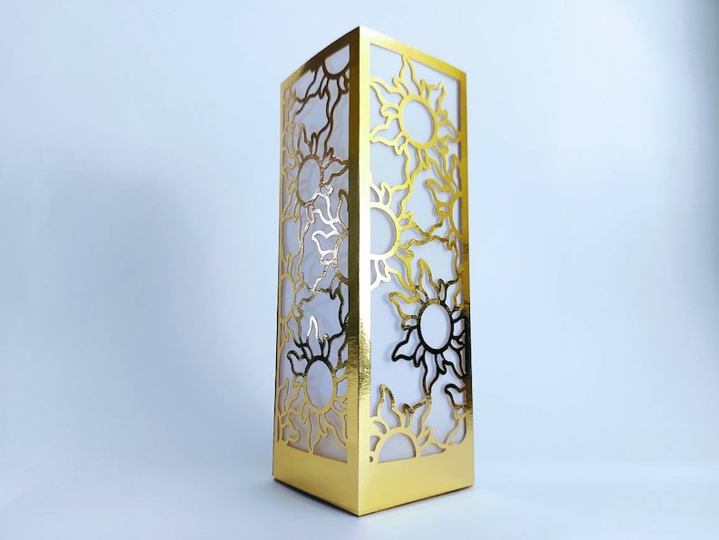 May include: A gold-colored decorative box with a cutout design of stylized suns. The box is made of cardboard and has a white interior.