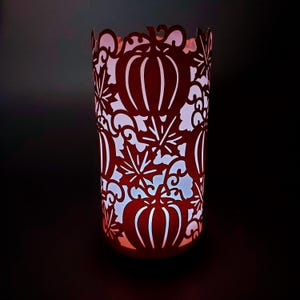 May include: A red metal candle holder with a cut-out design of pumpkins and leaves. The candle holder is cylindrical and has a scalloped top edge. The interior glows with a soft, white light, creating a warm ambiance. Perfect for fall decor.