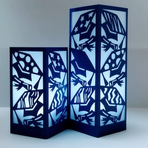 Puede incluir: Dos faroles de papel azules con intrincados diseños recortados. Los diseños presentan formas geométricas, estrellas y círculos. Los faroles están iluminados desde el interior, proyectando un suave resplandor.