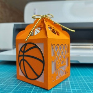 May include: Orange and black paper gift box with a basketball design and a gold ribbon. The box has a cut-out design of a basketball hoop and net.