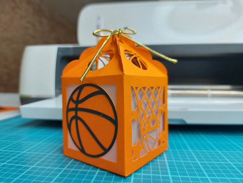 May include: Orange and black paper gift box with a basketball design and a gold ribbon. The box has a cut-out design of a basketball hoop and net.