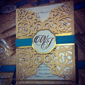 May include: Gold and teal wedding invitation with a decorative lace pattern and the initials "OWJ" in a circle. The text "Aliano Calab A las 09.00 pm Traje Formal V&aacute;lido para persona (s)" is printed on the invitation.