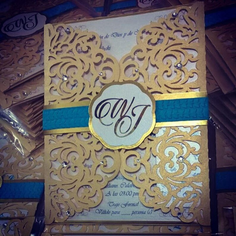 May include: Gold and teal wedding invitation with a decorative lace pattern and the initials "OWJ" in a circle. The text "Aliano Calab A las 09.00 pm Traje Formal V&aacute;lido para persona (s)" is printed on the invitation.