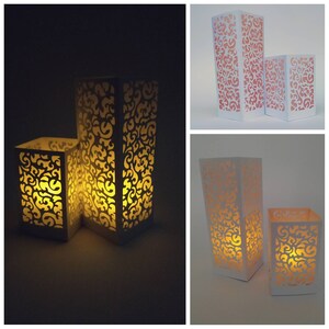 May include: Two white paper lanterns with intricate cut-out designs. The lanterns are illuminated from within, casting a warm glow. The smaller lantern is in front of the larger lantern.