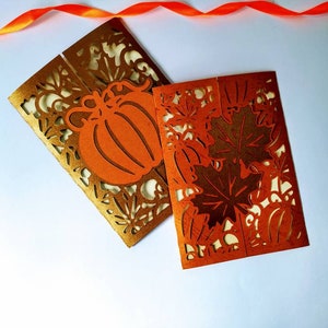 May include: Two folded cards with intricate cut-out designs. One card features a pumpkin and leaves in gold and orange, while the other card features leaves in orange and gold.