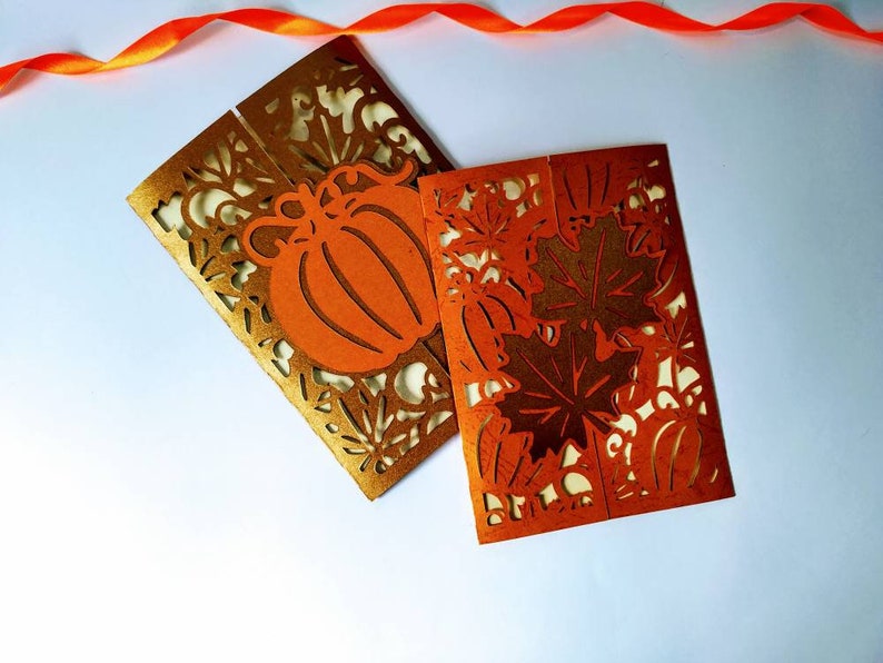 May include: Two folded cards with intricate cut-out designs. One card features a pumpkin and leaves in gold and orange, while the other card features leaves in orange and gold.