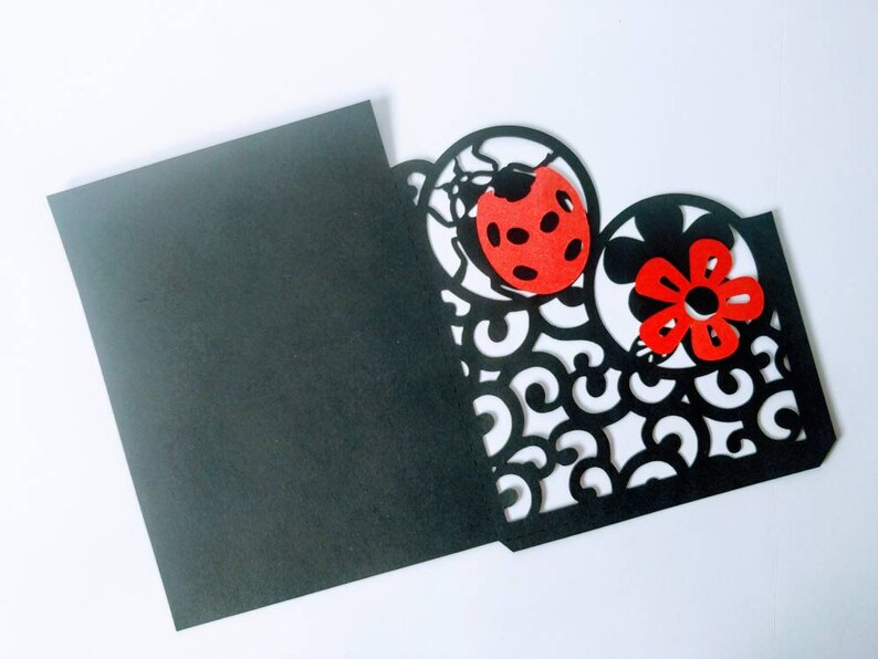 May include: Black card with a cut-out design of a ladybug and flower. The ladybug is red with black spots and the flower is red with a black center.
