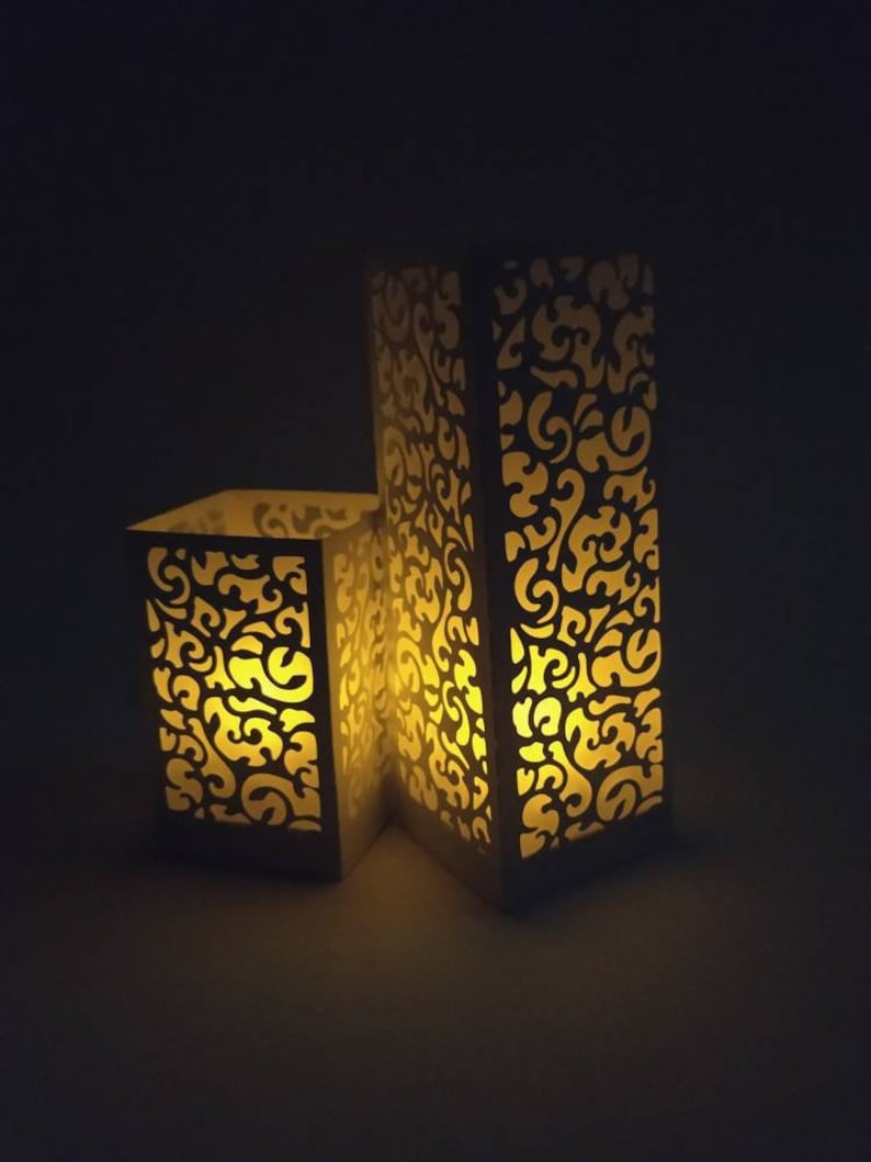 May include: Two white paper lanterns with intricate cut-out designs. The lanterns are illuminated from within, casting a warm glow.