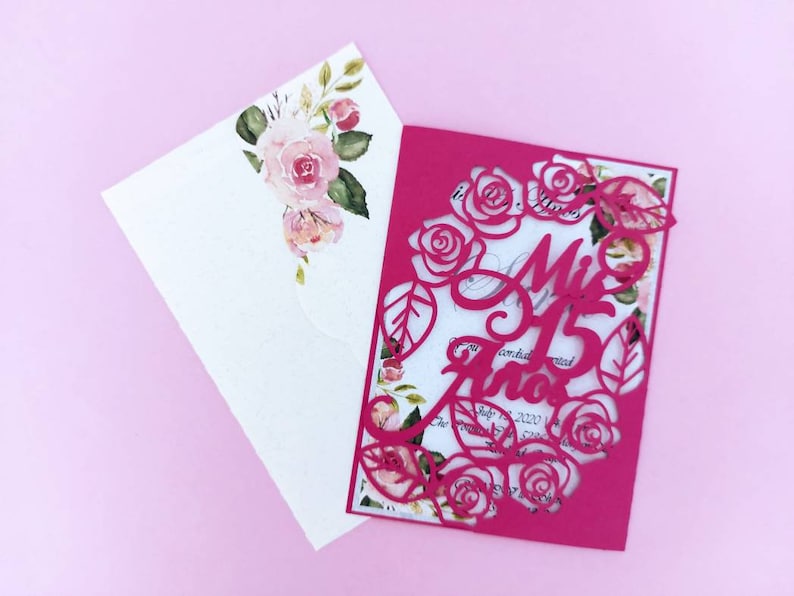 May include: A pink invitation with a cut-out design of roses and leaves. The text "Mis 15 Anos" is written in a decorative font.