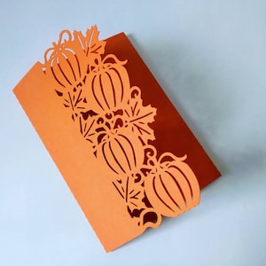 May include: Orange card with a decorative cut-out design of pumpkins and leaves. The card is folded in half.