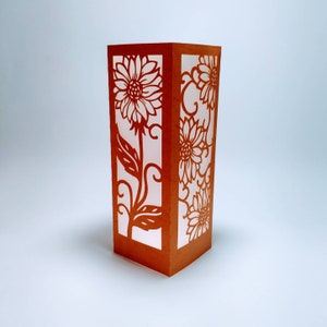 May include: Orange paper lantern with a white paper insert featuring a cut-out design of sunflowers and leaves.