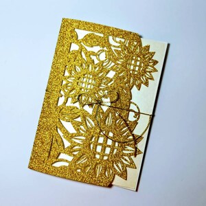 May include: Gold glitter laser cut wedding invitation with a sunflower design.