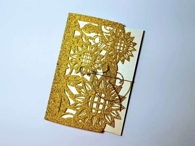 May include: Gold glitter laser cut wedding invitation with a sunflower design.