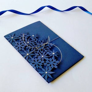 K&ouml;nnte beinhalten: Eine marineblaue Karte mit einem dekorativen Design aus blauen Papierblumen und silberner Schnur. Die Karte ist in der Mitte gefaltet und die Blumen befinden sich auf der Vorderseite der Karte.