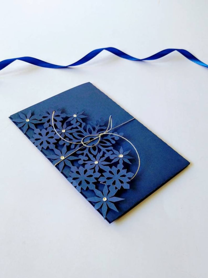 K&ouml;nnte beinhalten: Eine marineblaue Karte mit einem dekorativen Design aus blauen Papierblumen und silberner Schnur. Die Karte ist in der Mitte gefaltet und die Blumen befinden sich auf der Vorderseite der Karte.