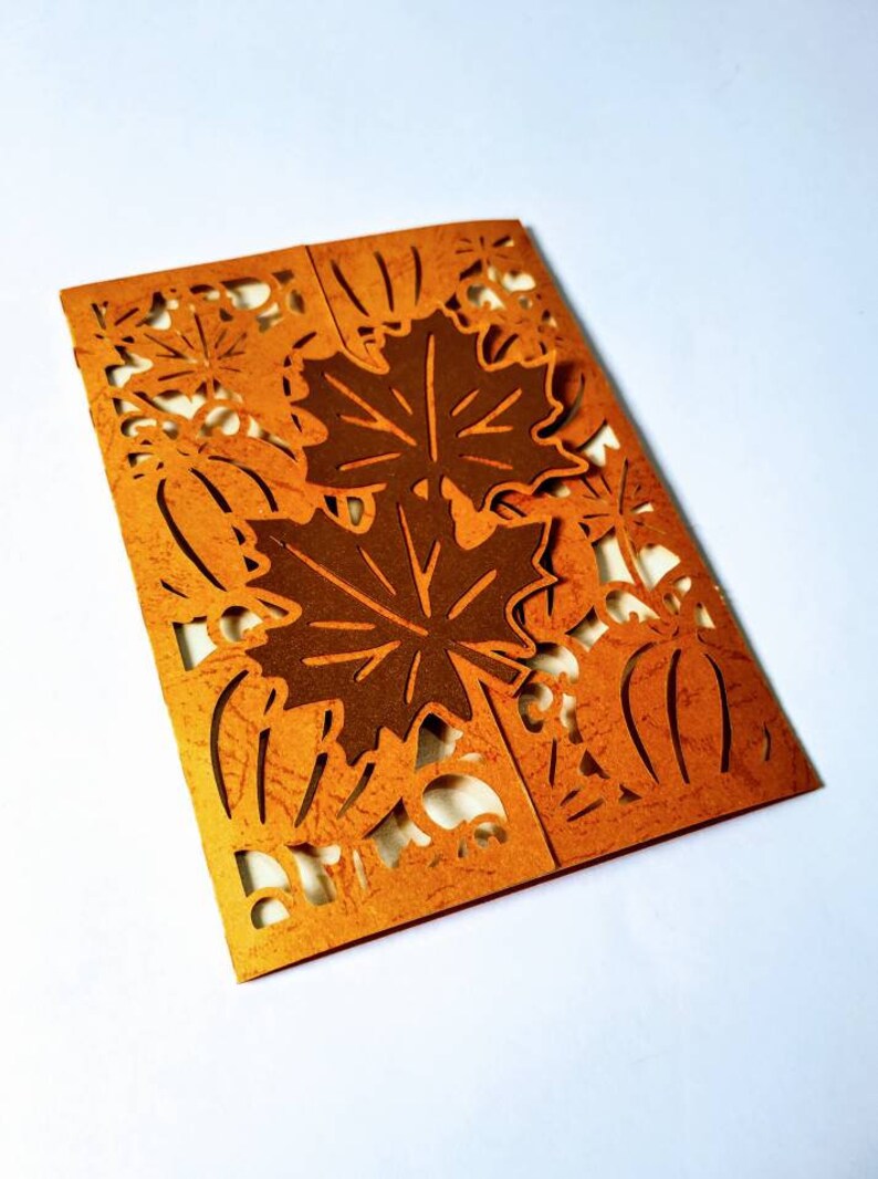May include: A brown card with a cut-out design of fall leaves and pumpkins. The card is folded in half and has a textured surface.