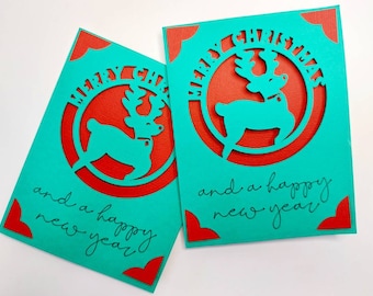 May include: Two turquoise Christmas cards with red accents. Each card features a reindeer cutout with the text "Merry Christmas and a happy new year" in black.