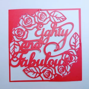 以下が含まれることがあります： 白い背景に赤い紙で切り抜かれたカード。カードには、バラと葉の円形の中に、「Eighty and Fabulous」という文字が装飾的なフォントで書かれている。