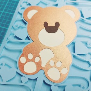 May include: A light blue card with a brown teddy bear cutout. The teddy bear has white paws and a white face with brown ears. The card is decorated with a blue and white swirl design.