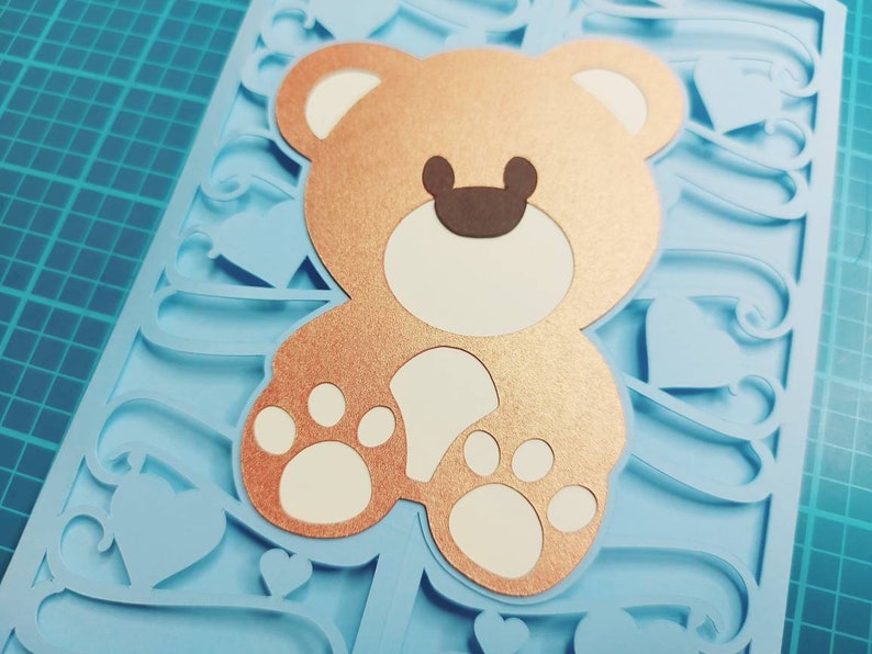 May include: A light blue card with a brown teddy bear cutout. The teddy bear has white paws and a white face with brown ears. The card is decorated with a blue and white swirl design.