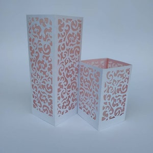 May include: Two white paper lanterns with intricate cut-out designs. The lanterns are made of white paper with a pink floral pattern. The larger lantern is taller than the smaller lantern.