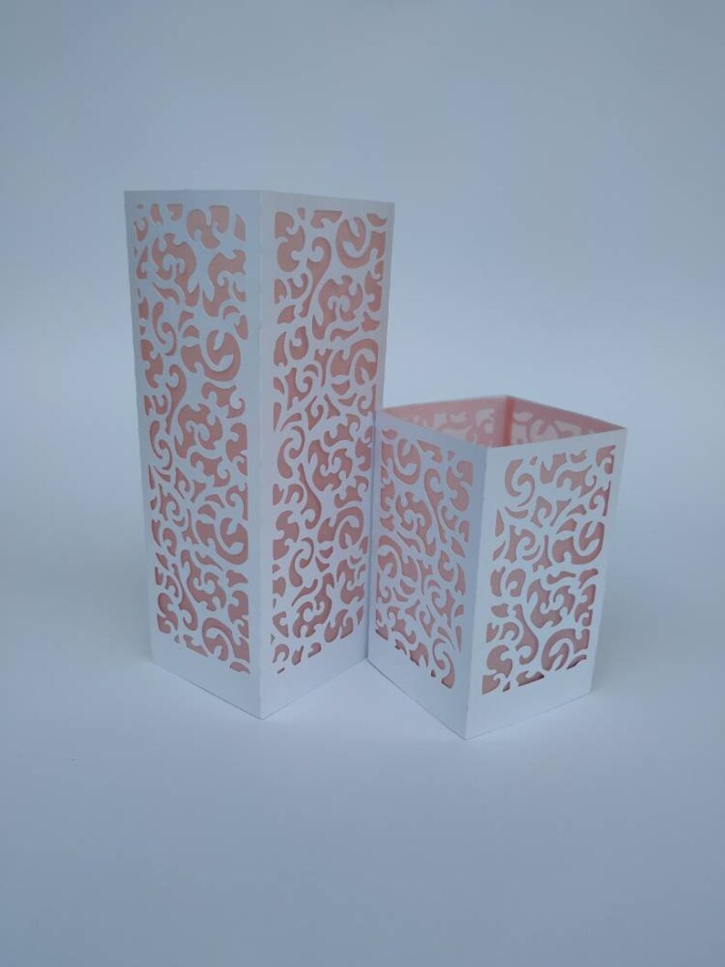 May include: Two white paper lanterns with intricate cut-out designs. The lanterns are made of white paper with a pink floral pattern. The larger lantern is taller than the smaller lantern.