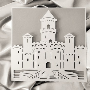 May include: A white paper cut-out of a castle with a tall central tower and four smaller towers. The castle has a drawbridge and a gate.