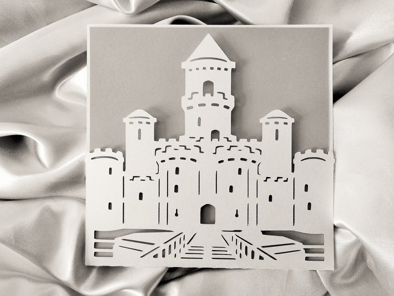 May include: A white paper cut-out of a castle with a tall central tower and four smaller towers. The castle has a drawbridge and a gate.