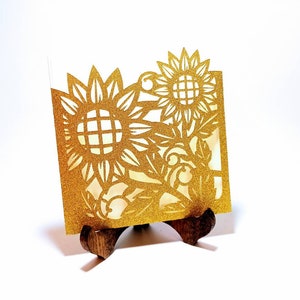 May include: Gold glitter card with a cut-out design of sunflowers and leaves. The card is displayed on a small wooden stand.
