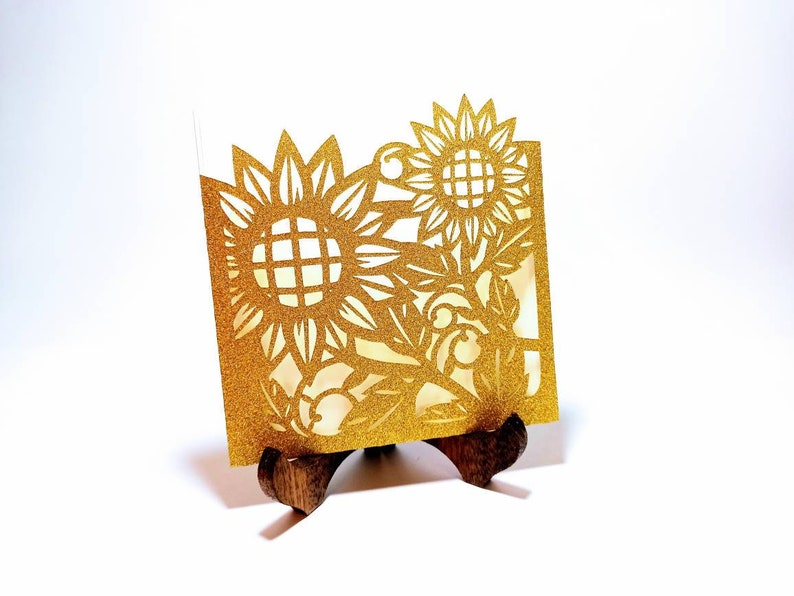 May include: Gold glitter card with a cut-out design of sunflowers and leaves. The card is displayed on a small wooden stand.