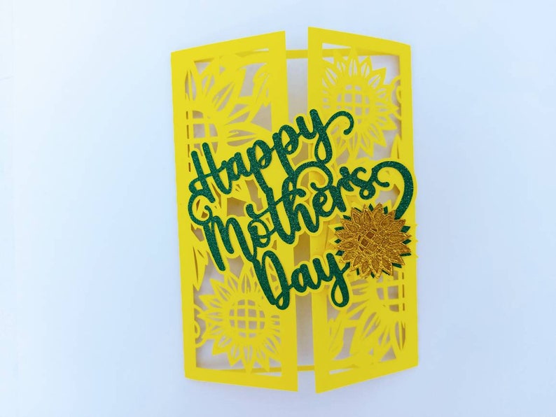 Op de afbeelding: Een gele kaart met een groene glitter-uitsparing van de woorden "Happy Mother's Day" en een gouden glitter-zonnebloem. De kaart is versierd met een zonnebloemmotief.