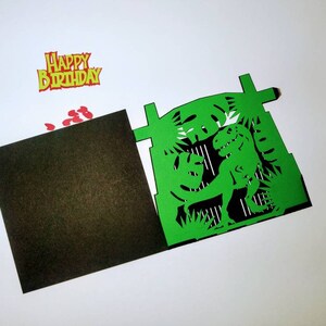 May include: Green paper card with a pop-up dinosaur inside. The dinosaur is green and has a black outline. The card is folded in half and has a black inside. The card has a red and yellow "Happy Birthday" banner on top.