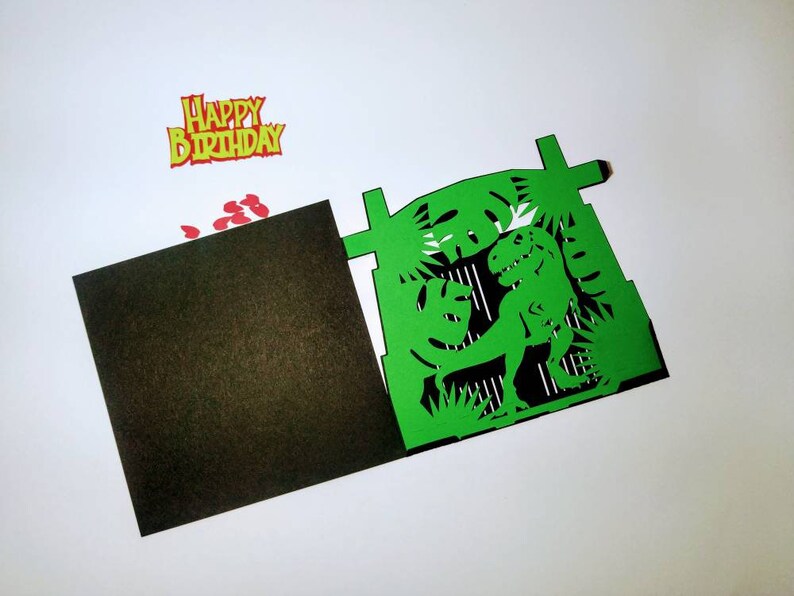 May include: Green paper card with a pop-up dinosaur inside. The dinosaur is green and has a black outline. The card is folded in half and has a black inside. The card has a red and yellow "Happy Birthday" banner on top.
