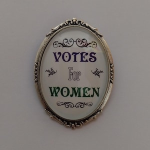 May include: A silver oval-shaped pin with a white background and the text "Votes For Women" in green and purple lettering.