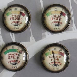 May include: Four round, brass-colored gauges with glass faces. Each gauge has a different colored needle and a different label: Steam Pressure, Field Density, Power Level, and Radiation μw/h. Each gauge has the text "K. Beggart Ltd" and "Socr" printed on the face.